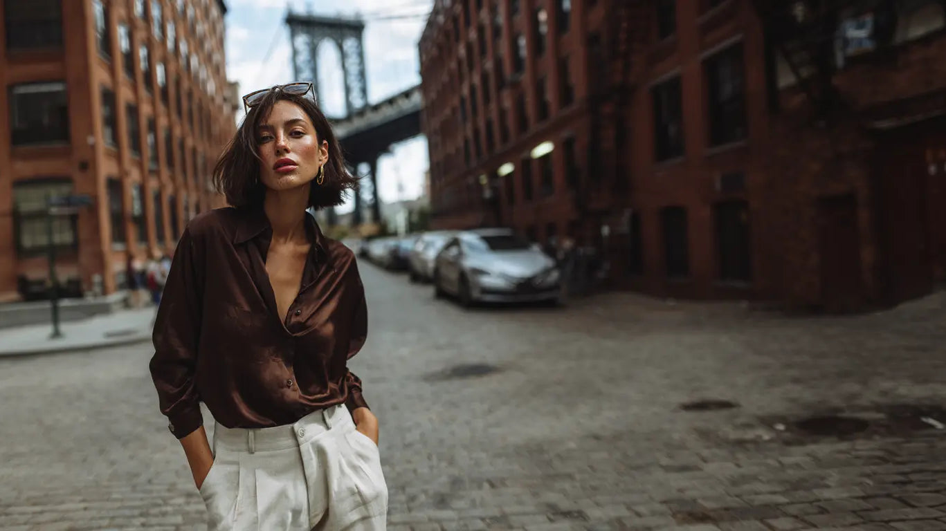 Tuck-Guide Editorial: Frau in dunkelbrauner Seidenbluse und cremefarbener Hose vor Manhattan Bridge in DUMBO Brooklyn.