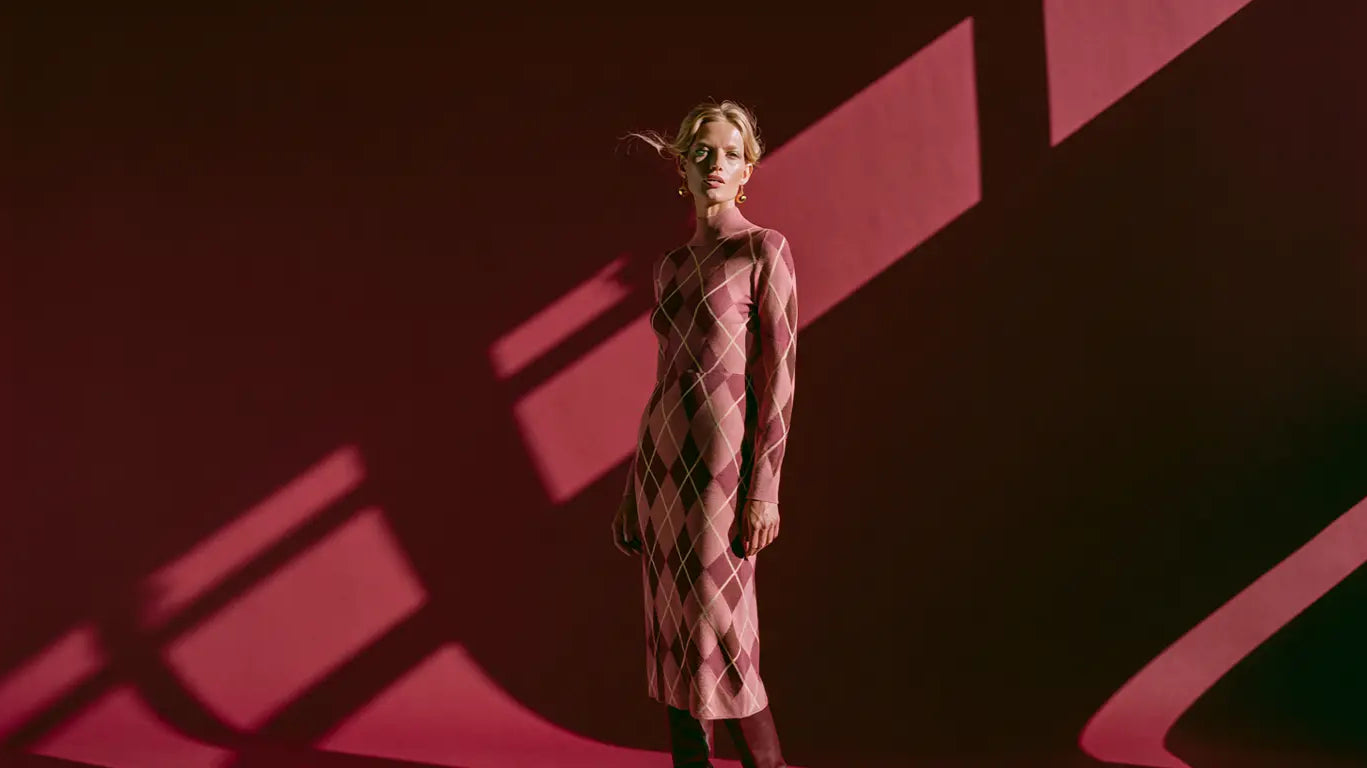 Frau im taillierten Argyle-Strickkleid in Blush, Mauve und Braun vor tiefrotem Hintergrund – KI-Illustration