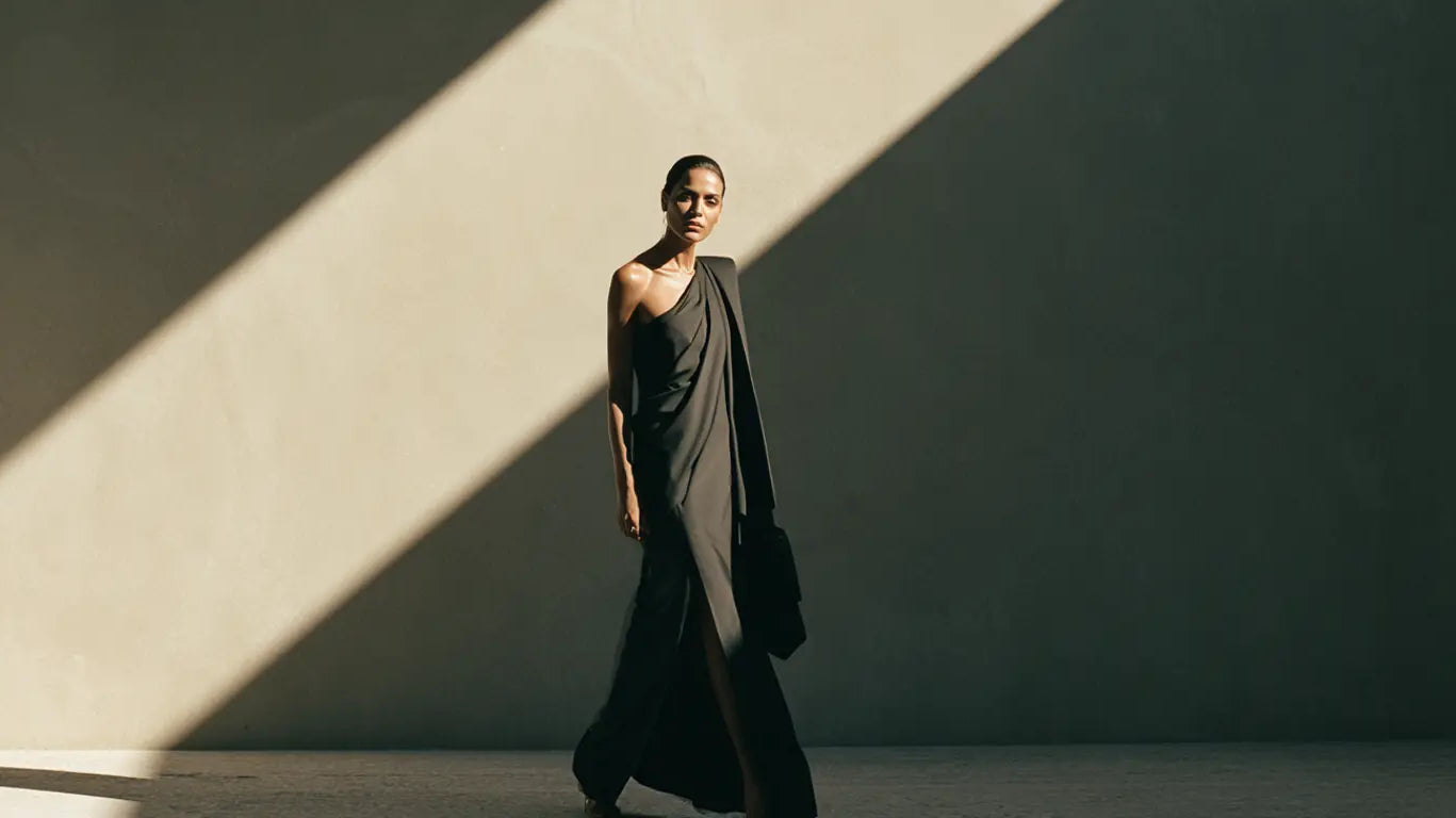 Frau im schwarzen asymmetrischen One-Shoulder-Kleid mit einer freien Schulter vor minimalistischer Betonwand mit diagonalem Lichtspiel – KI-Illustration