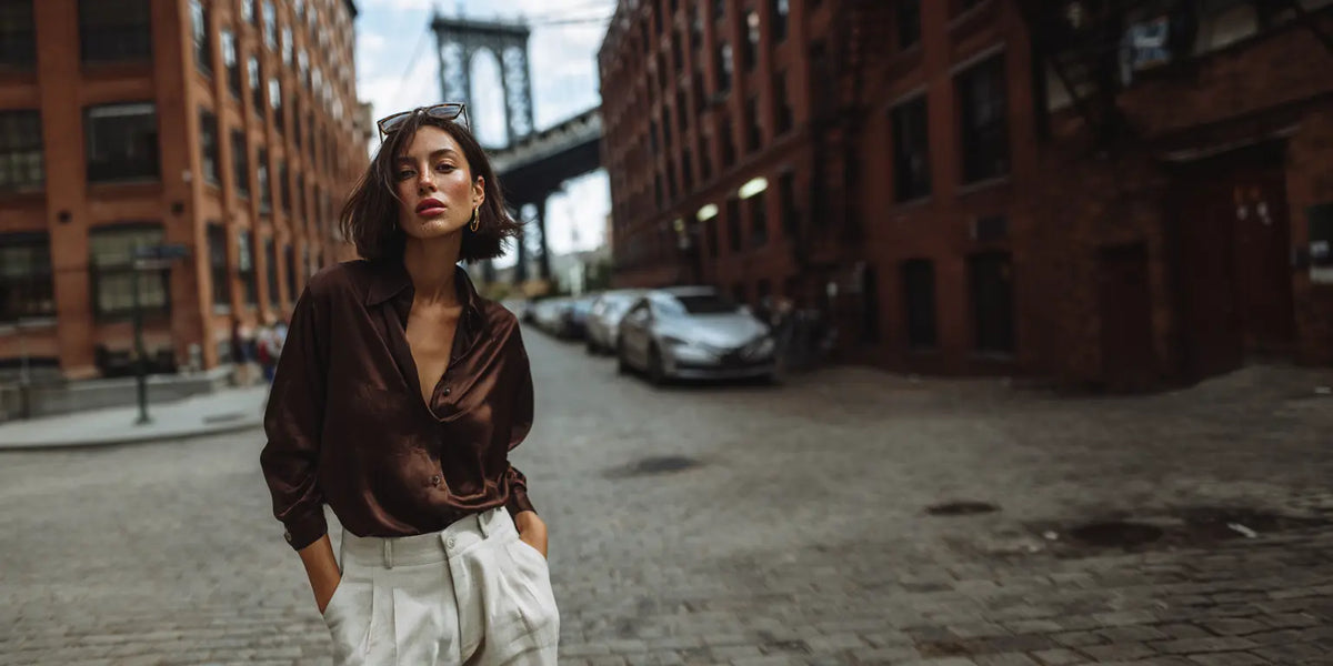 Tuck-Guide Editorial: Frau in dunkelbrauner Seidenbluse und cremefarbener Hose vor Manhattan Bridge in DUMBO Brooklyn.
