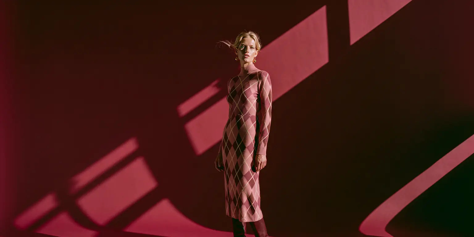 Frau im taillierten Argyle-Strickkleid in Blush, Mauve und Braun vor tiefrotem Hintergrund – KI-Illustration