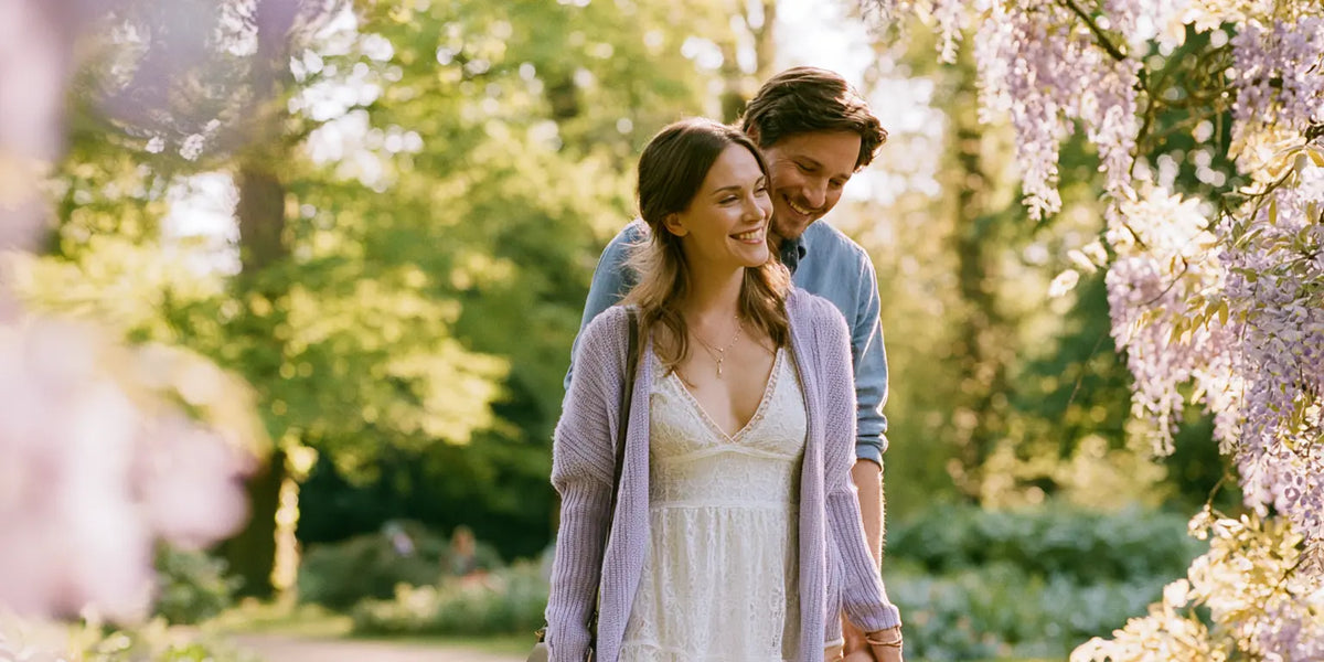 Braut im weißen Spitzenkleid mit fliederfarbenem Strickcardigan, Hand in Hand mit ihrem Partner in einem sonnigen Park voller Glyzinien – KI-Illustration
