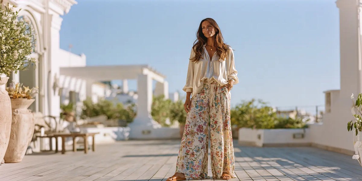 Frau in floral gemusterter weiter Sommerhose mit oversized cremefarbener Satinbluse auf einer sonnigen mediterranen Terrasse – KI-Illustration